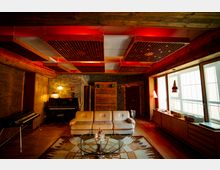 Gemütlicher Raum im Retro-Stil mit hölzernen Wänden, einer beigefarbenen Couch, einem Glastisch und einem Teppich in geometrischem Muster. Die Akustikdecke mit roten Elementen, ein Klavier, sowie große Fenster mit einfallendem Tageslicht prägen die Atmosphäre.