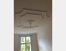 Zimmerdecke mit dekorativem Stuckrahmen und einer Rosette in der Mitte, aus der ein Kabel für eine Lampe herunterhängt. Im Hintergrund ist ein großes Fenster mit weißem Rahmen und Blick auf grüne Bäume zu sehen.