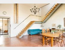 Großzügiger Wohnraum mit einer Holztreppe in modernem Design, die zu einer offenen Empore führt. Im Raum befinden sich ein blauer Sofa, ein runder Holztisch mit mehreren Stühlen, ein Kronleuchter sowie dekorative Elemente wie Bilder und Pflanzen auf Möbeln.