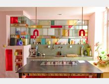 Bright modern kitchen with a large central metal island and built-in stovetop, backed by gray tile counters and shelves lined with bottles and small appliances. A wall of colorful stained-glass-style panels and two red U-shaped pendant lights frame the space, with sunlight coming in from tall windows on the right.