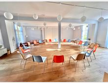 Großer, heller Seminarraum mit Holzboden, in dem Stühle in einem großen Kreis aufgestellt sind. Von der Decke hängen dekorative, runde Lampen, und große Fenster lassen viel Tageslicht herein.