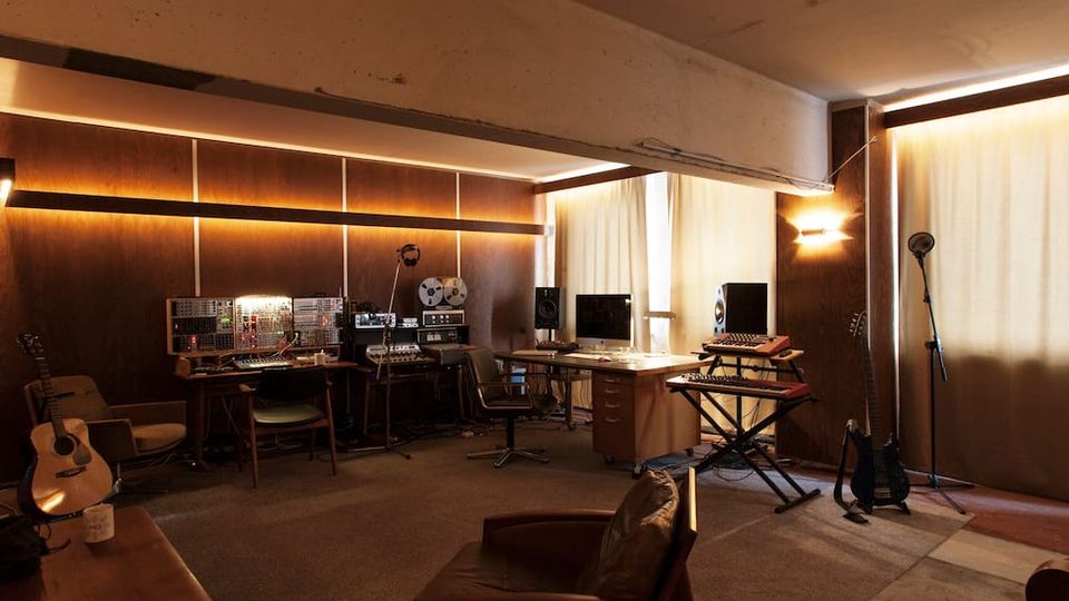 Innenraum eines Tonstudios mit Holzvertäfelung und warmen Wandleuchten, ausgestattet mit Mischpulten, Aufnahmegeräten und Monitoren an mehreren Arbeitsplätzen. Große, mit Vorhängen bedeckte Fenster sowie Musikinstrumente wie Gitarren, Keyboard und Mikrofonständer prägen den Raum.