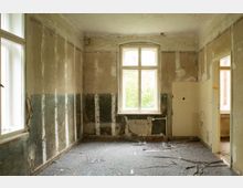 Leerer, entkernter Innenraum mit abgenutzten, teils abgeplatzten Wänden und beschädigtem Bodenbelag. Zwei hohe Fenster und ein Türdurchgang lassen Tageslicht herein; unter dem mittleren Fenster sind alte Heizungsrohre sichtbar.