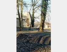 Ein Park mit kahlen Bäumen im Winter, mit einem gewundenen Gehweg, der durch das Gelände führt. Im Hintergrund ist ein historisches Gebäude mit heller Fassade und einem Türmchen sichtbar.