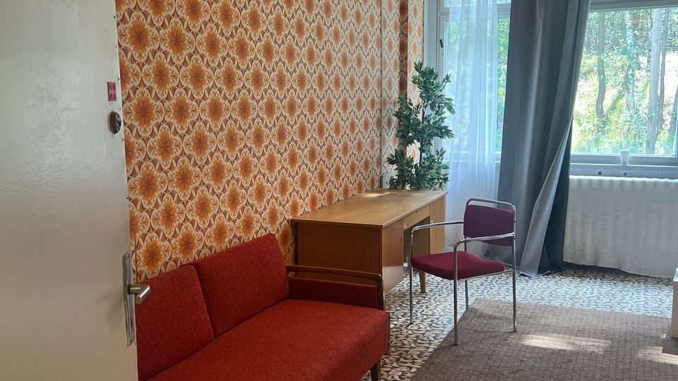 Kleines Wohnzimmer mit orange gemusterter Tapete, rotem Sofa, kleinem Holztisch und Stuhl sowie gemustertem Fliesenboden mit Teppich. Am Fenster mit Vorhängen steht eine Pflanze; an der Decke hängt eine große, runde Deckenlampe.