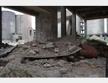 Industriegelände mit verlassenen Gebäuden und zerstörten Strukturen. Im Vordergrund ein Haufen aus Bauschutt und Trümmern, im Hintergrund betonierte Silos und ein gemauerter Schornstein.