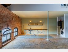 Loftartige Küche mit Backsteinwand und zwei bogenförmigen Fenstern, einer freistehenden Kücheninsel aus Metall und Holz, und Regalen mit Geschirr an der hellgrünen Wand. Rechts befinden sich ein Einbauschrank mit Mikrowelle und eine Waschmaschine.