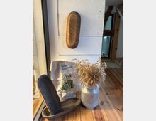 Ein rustikales Arrangement in einem Innenraum mit Holzboden: ein hängender und ein liegender Brotkorb aus Holz, ein Sack Roggenmehl mit Aufschrift „Bestes Roggenmehl“ und ein Gefäß aus Metall mit darin stehenden Ähren. Im Hintergrund sind weiße Holzwände und sichtbare Holzbalken zu sehen.