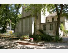 Außenbereich eines Gebäudes mit grobem, beige-grünem Putz, umgeben von Bäumen. Im Vordergrund stehen Bierbänke, Blumenkübel, eine Rampe, Treppenstufen und ein Geländer; Girlanden mit Lichterketten spannen sich über den Hof.