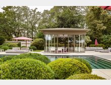 Moderne Gartenlandschaft mit einem gläsernen Pavillon im Zentrum, umgeben von gepflegten Büschen, einem Pool und Sitzbereichen mit Sonnenliegen und roten Sonnenschirmen. Im Hintergrund befinden sich große Bäume und dichtes Grün.