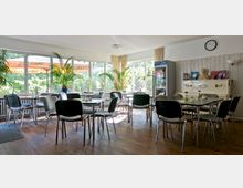 Heller Aufenthaltsraum mit mehreren kleinen Tischen und grauen Stühlen, großen Fenstern mit Blick ins Grüne, dekorativen Pflanzen und einem weißen Klavier an der Wand. Im Hintergrund ein Kühlschrank mit Getränken und Prospekten auf einem Regal.