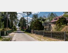 Ruhige Wohnstraße mit freistehenden Häusern, umgeben von hohen Bäumen und gepflegten Gärten. Die Straße ist asphaltiert und von schmiedeeisernen Zäunen sowie Hecken gesäumt.