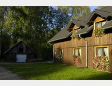 Holzhaus mit dunkelgrauem Ziegeldach und Dachgauben, dessen Fenster mit Blumenkästen dekoriert sind. Im Hintergrund steht ein kleineres Holzgebäude mit Schrägdach, umgeben von einem Garten und hohen Bäumen.