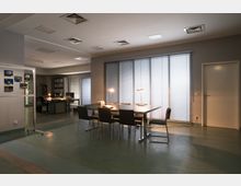 Büroinnenraum mit einem großen Konferenztisch und mehreren Stühlen in der Mitte, beleuchtet von Tischlampen. Im Hintergrund ein Schreibtisch mit Computer und Aktenschrank sowie eine Wand mit Lamellenjalousien vor Fenstern.