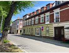 Straßenszene mit historischen Wohn- und Geschäftshäusern, teils mit rot-brauner Backsteinfassade und grünen Farbakzenten; gepflasterter Gehweg und eine Baumreihe säumen die asphaltierte Straße, die unter einem blauen Himmel verläuft.