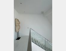 Moderner, heller Innenraum mit einer weißen Wand, an der eine skulpturale Figur auf einem schwarzen Ständer lehnt. Rechts im Bild sind ein Treppengeländer aus Glas und Metall sowie eine Treppe darunter zu sehen.