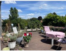 Große Holzterrasse mit Topfpflanzen, darunter blühende rote und grüne Pflanzen, sowie Gartenmöbeln wie einem runden Tisch mit rosa Tischdecke und Liegestühlen; im Hintergrund dichte grüne Büsche und Bäume unter blauem Himmel mit weißen Wolken.