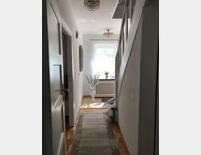 Ein heller Flur mit Holzboden und einem langen Teppichläufer. An der rechten Seite führt eine Treppe nach oben, geradeaus steht eine Vase mit Blumen vor einem Fenster mit weißem Rahmen und einem Heizkörper darunter, beleuchtet von natürlichem Licht.