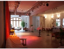 Ein Veranstaltungsraum mit hoher Decke, dekoriert mit Lichterketten und roten Vorhängen. Der Raum enthält kleine Stehtische mit weißen Tischdecken, Sitzgruppen aus Rattanmöbeln und große Fenster, die Tageslicht hereinlassen; Pflanzen und dezente Beleuchtung ergänzen das Ambiente.