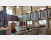 Innenansicht einer historischen Kirche mit bemaltem Holzgestühl und einer Empore mit blauen Paneelen und goldenen Zierleisten. Links steht ein reich verzierter Kanzelaltar mit Schnitzereien, unten ist ein Tisch mit Altardecke, Bibel und Blumenschmuck zu sehen.