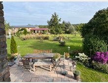 Großer Garten mit einer gepflasterten Terrasse im Vordergrund, auf der ein Holztisch mit Stühlen und Blumendekoration steht. Im Hintergrund sind grüne Wiesen, Bäume, Blumenbeete und ein Gebäude mit roten Dachziegeln zu sehen.