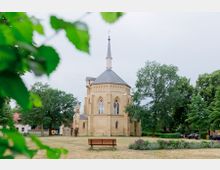 Eine kleine Kirche aus gelbem Backstein mit spitzem Turm und gotischen Fenstern, umgeben von Bäumen und einer Grünfläche. Im Vordergrund steht eine Holzbank, umgeben von Blumenbeeten.