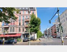 Straßenszene in einer urbanen Umgebung mit historischen Wohngebäuden, darunter ein rosa Gebäude mit weißen Verzierungen und einem roten Vordach. Im Vordergrund befindet sich eine Ampel, während Autos und Fußgänger auf der gepflasterten Straße zu sehen sind; ein Straßenschild weist auf die "Reichenberger Str." hin.