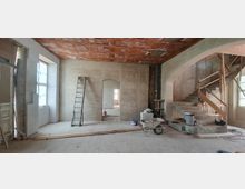 Innenraum eines Gebäudes in Renovierung mit unverputzten Wänden und einer unverkleideten Decke aus Ziegeln. Zu sehen sind eine Leiter, Baumaterialien, ein Schubkarren und eine provisorische Treppe aus Beton sowie ein Durchgang, der in einen weiteren Raum führt.