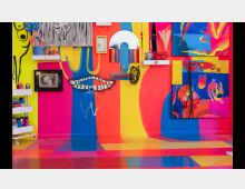 Wand eines kreativen Ateliers mit leuchtend bunten Farben in Pink, Gelb und Blau, dekoriert mit Kunstwerken, Regalen und Malutensilien. Der Boden zeigt ebenfalls ein farbenfrohes Muster, passend zur Gestaltung der Wände.