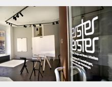 Ein heller Raum mit großen Fenstern, ausgestattet mit zwei weißen Tafeln, schwarzen Hockern und Schienenleuchten an der Decke. Im Vordergrund ist eine Glastür mit dem Schriftzug "ester erster", der auf Kunst, Design und Vermietungen hinweist.
