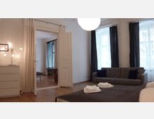 Helles Wohnzimmer mit Holzboden, grauem Sofa und großen Fenstern mit weißen und dunklen Vorhängen. Im Vordergrund ein Bett mit gefalteten Handtüchern, daneben ein weißer Schrank mit einer Lampe, im Hintergrund ein angrenzender Raum mit geöffneten Doppeltüren.