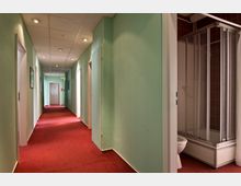 Ein Innenraum mit einem langen, hell beleuchteten Flur, grün gestrichenen Wänden und rotem Teppichboden. Rechts im Vordergrund befindet sich ein offenstehendes Badezimmer mit einer Dusche und einer Toilette.