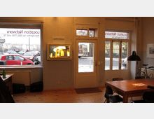 Innenraum eines Büros mit großen Fenstern und Glastür, die auf eine Straße blicken. Im Raum befinden sich ein Holztisch mit Stühlen, eine Wandvitrine mit Beleuchtung, sowie Fahrräder und Bürozubehör.