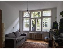 Ein Wohnzimmer mit einem großen Fenster, das Blick auf einen Baum und ein gegenüberliegendes Gebäude bietet. Im Raum stehen ein graues Sofa, ein Teppich auf Holzboden, DJ-Equipment auf einem Regal und Zimmerpflanzen auf Möbeln; ein Heizkörper ist unter dem Fenster installiert.