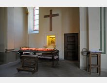 Innenraum einer Kirche mit einem Bereich für das Entzünden von Kerzen. Zu sehen sind ein Holzkreuz mit der Aufschrift „Heilige Mission“, ein beleuchtetes Marienbild, zahlreiche brennende Kerzen auf einem Metallgestell, Blumenvasen sowie eine Kniebank und ein Beistelltisch.