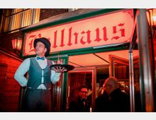 Ein Eingang zu einem Veranstaltungsort mit dem Schriftzug „Ballhaus“ in roten Buchstaben auf einem beleuchteten Schild. Im Vordergrund steht ein Kellner in Vintage-Kleidung mit Tablett, daneben sind weitere Personen unter einem Vordach zu sehen.