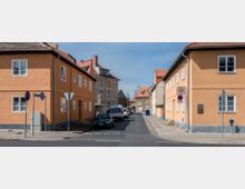 Straßenansicht einer Wohnsiedlung mit orangefarbenen verputzten Gebäuden, roten Ziegeldächern und weißen Fenstern. Parkende Autos säumen die schmale Straße, und Verkehrsschilder weisen auf eine Tempo-30-Zone hin.