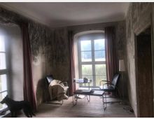 Ein schlichtes Zimmer mit zwei großen Fenstern, die mit roten Vorhängen dekoriert sind, und abgenutzten Wänden mit sichtbaren Spuren von Feuchtigkeit. Der Raum enthält zwei schwarze Sessel, einen kleinen Glastisch, eine Stehlampe und einen Holzboden; ein Hund steht am Fenster.