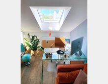 Ein helles Dachzimmer mit einem modernen Arbeitsplatz. Zu sehen sind ein Glastisch mit Computer und Büromaterial, ein brauner Drehstuhl, eine große Zimmerpflanze, ein Globus, ein Dachfenster mit Blick nach draußen und ein orangefarbener Sessel.