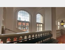 Innenansicht eines historischen Treppenhauses mit großen, gebogenen Fenstern und einer weißen Balustrade. Im Hintergrund ist eine dekorative Laterne zu sehen; durch die Fenster fällt Tageslicht, das den Raum erhellt.
