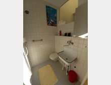 Kleines Badezimmer mit weiß gefliesten Wänden und grauem Fliesenboden. Ein Waschbecken mit Wandarmatur, eine Toilette, ein Handtuchhalter sowie eine gelbe Badematte sind zu sehen, dazu ein roter Eimer und eine Wanddekoration mit einem Unterwasser-Motiv.