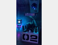 Ein futuristischer Gaming-Raum mit einer Tischreihe voller Computer, Tastaturen und Headsets, beleuchtet von blauem und violettem Licht. Im Hintergrund befinden sich ein großformatiger Lüfter an der Decke und ein automatisches Gerät mit einem Bildschirm, das an eine digitale Steuereinheit erinnert.