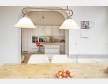 Helle, moderne Wohnküche mit weißer Einbauküche, zentraler Kücheninsel und rot gepolstertem Barhocker. Im Vordergrund ein Holztisch mit Schale voller Äpfel und darüber zwei hängende Deckenlampen.