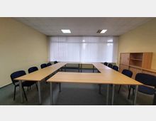 Ein einfacher Konferenzraum mit U-förmig angeordneten Holztischen und dunkelblauen Stühlen. Im Hintergrund befinden sich große Fenster mit vertikalen Jalousien und rechts ein Holzregal mit Schränken.