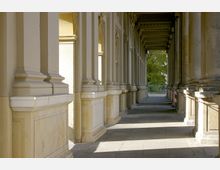 Blick in einen klassischen Säulengang mit hellen Steinpfeilern, dekorativen Kapitellen und einem überdachten, schattigen Gehweg. Im Hintergrund ist eine Grünfläche mit Bäumen erkennbar.