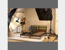 Eine Fotostudio-Szene mit einem Bettgestell im Vordergrund, umgeben von Beleuchtungsausrüstung, darunter zwei große Softboxen. Im Hintergrund ist eine Backsteinwand sichtbar, und eine Person richtet das Set aus.