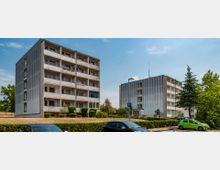 Zwei vierstöckige Wohngebäude mit klaren, rechteckigen Fassaden und Balkonen, umgeben von gepflasterten Wegen und kleinen Grünflächen mit Bäumen und Büschen. Im Vordergrund ist ein Parkplatz mit geparkten Autos zu sehen.