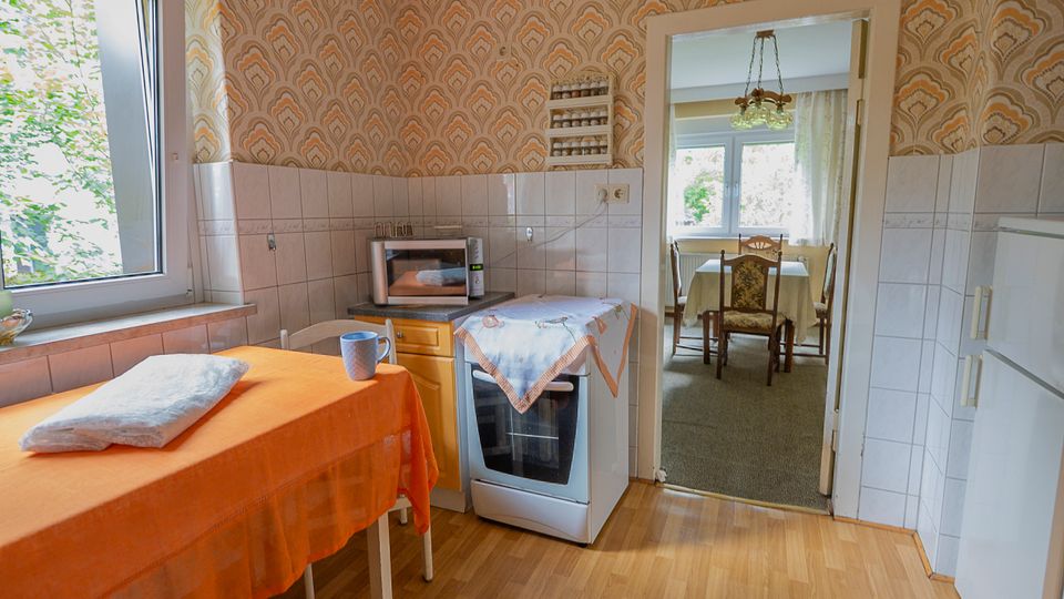 Blick in eine kleine Küche mit großem Fenster, gemusterten Tapeten und halbhoch gefliesten Wänden, mit Tisch, Herd, Mikrowelle und Kühlschrank. Durch eine offene Tür ist ein angrenzendes Esszimmer mit Tisch, Stühlen und Hängelampe zu sehen.