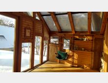 Holzveranda mit großen Glasfenstern und einem schrägen Glasdach, die viel Tageslicht hereinlassen. Im Raum befinden sich ein Hängesessel, Holzwände mit Regalen und Blick auf eine verschneite Außenlandschaft.