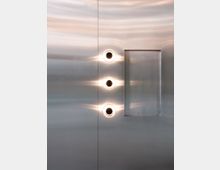 Metallische Wand mit drei runden, leuchtenden Öffnungen in einer vertikalen Linie und einem rechteckigen Ausschnitt rechts daneben, reflektierend und minimalistisch gestaltet.